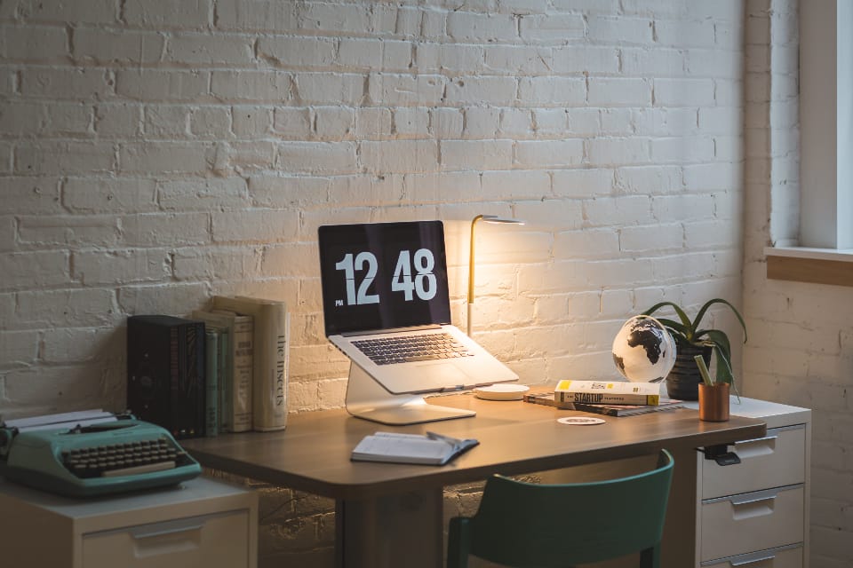 An office desk with a laptop, books and a typewriter against a brick wall